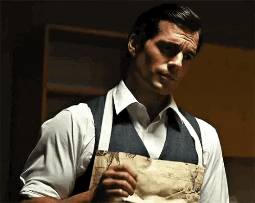a man wearing an apron and vest is standing in a kitchen holding a piece of paper .