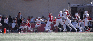 a group of football players are running on a field with a referee watching .