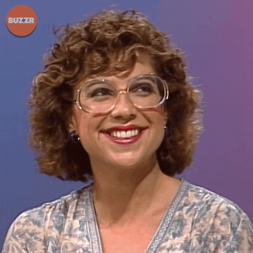 a woman with curly hair and glasses is smiling in front of a buzzr button