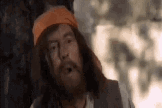 a man with long hair and a beard is wearing an orange headband and a vest .