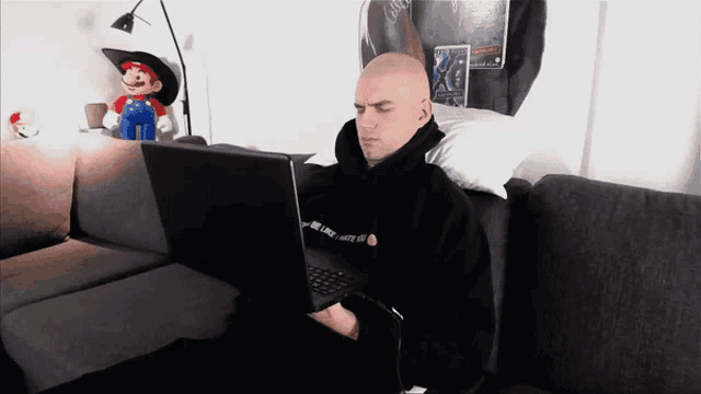 a bald man sits on a couch using a laptop with a mario doll behind him
