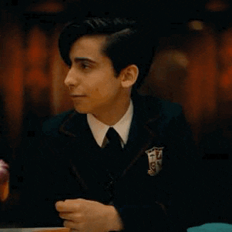 a young man in a suit and tie is sitting at a table with his hands out .