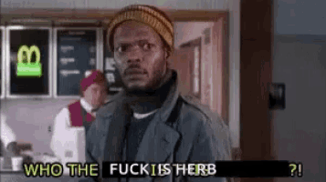 a man in a hat and coat is standing in front of a sign that says ' who the fuck is herb ? '