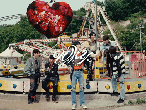 a group of young men are standing in front of a roller coaster with a heart shaped balloon in the background that says we