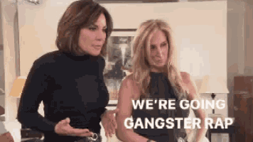 two women are standing next to each other in a living room and one of them says we 're going gangster rap .