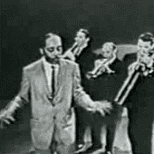 a black and white photo of a man in a suit and tie standing in front of a group of men playing instruments .