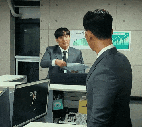 a man in a suit and tie is talking to another man in front of a computer monitor