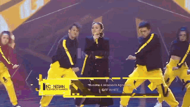 a woman singing into a microphone with a group of dancers in yellow