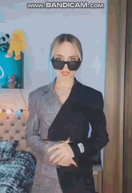 a woman wearing sunglasses and a suit is standing in a bedroom .