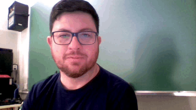 a man with glasses and a beard stands in front of a green board