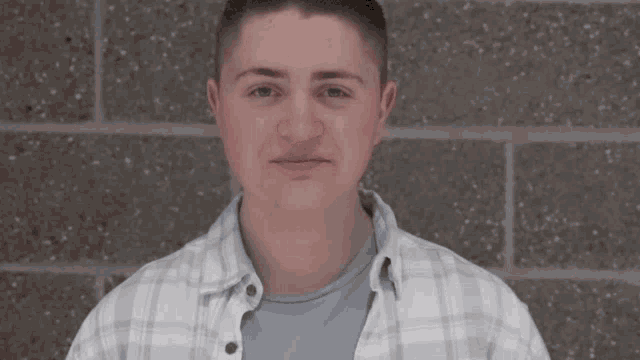 a young man standing in front of a brick wall wearing a plaid shirt