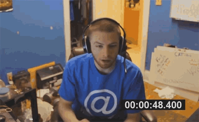 a man wearing headphones and a blue shirt with an @ on it is sitting at a desk