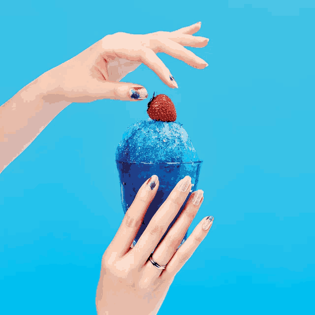 a woman 's hand reaches for a strawberry on top of a blue shaved ice dessert