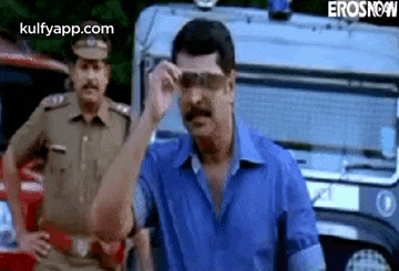 a man wearing sunglasses is standing in front of a police car while a police officer stands behind him .