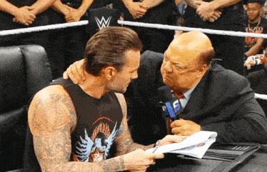 a man with a tattoo on his arm talks to another man in a wrestling ring