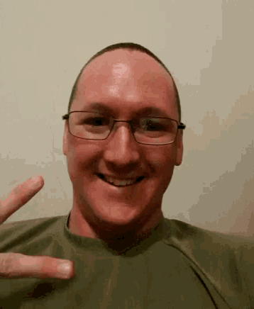 a man wearing glasses is smiling and making a peace sign with his hand .