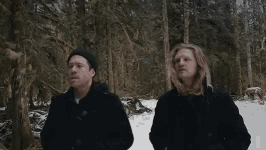 two men are standing in the snow in the woods looking at something .