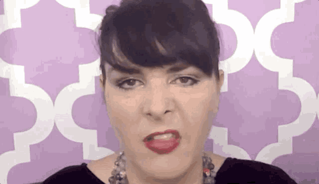 a woman with black hair and red lipstick is making a funny face in front of a purple background .