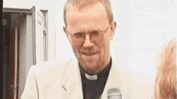 a priest wearing glasses and a white jacket is holding a microphone .
