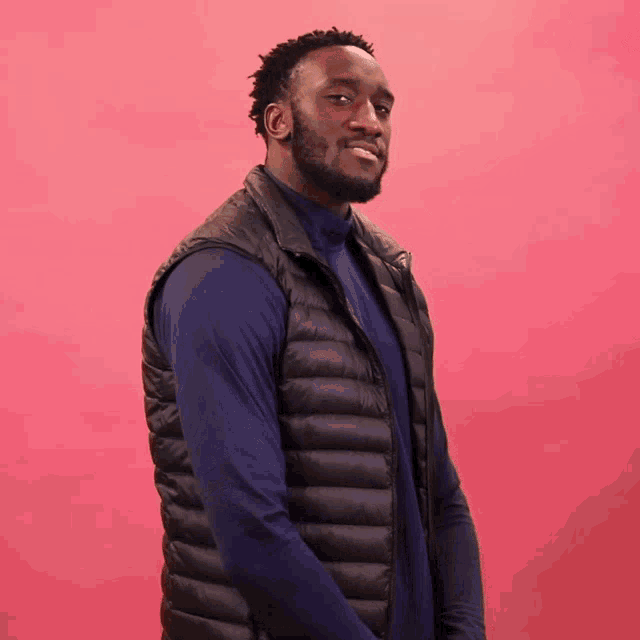a man wearing a black vest and blue shirt is standing in front of a pink background