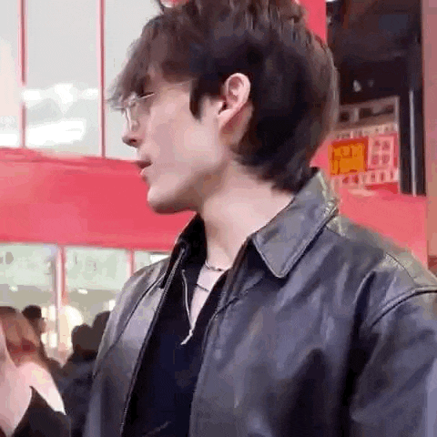 a young man wearing glasses and a black leather jacket is standing in front of a red building .