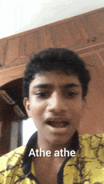 a young man wearing a yellow shirt says ' athe athe ' in front of a wooden wall