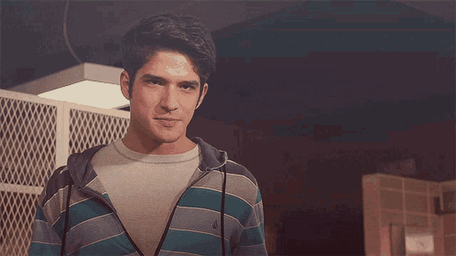 a young man wearing a striped shirt and a hoodie stands in front of a chain link fence