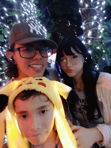 a man wearing a ny hat stands next to a girl wearing glasses and a boy wearing a pikachu headband