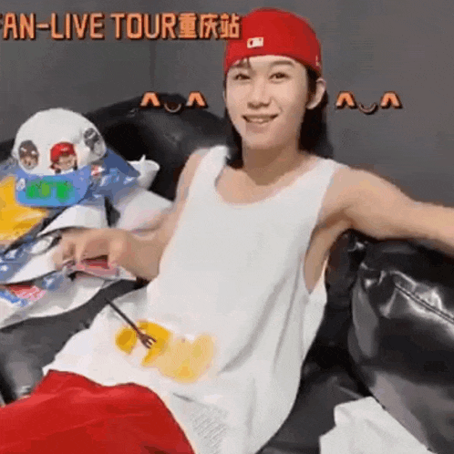 a man wearing a red hat and a white tank top is sitting on a couch eating food .
