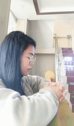 a woman wearing glasses sits with her hands folded in front of a set of stairs