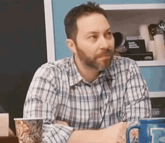 a man in a plaid shirt is sitting at a table with his arms crossed and a cup of coffee .
