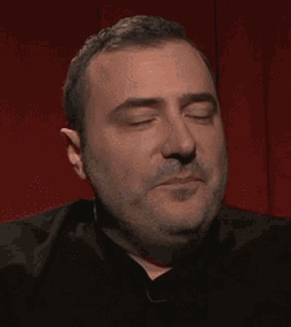 a man with a beard is sitting in front of a red curtain and making a face .