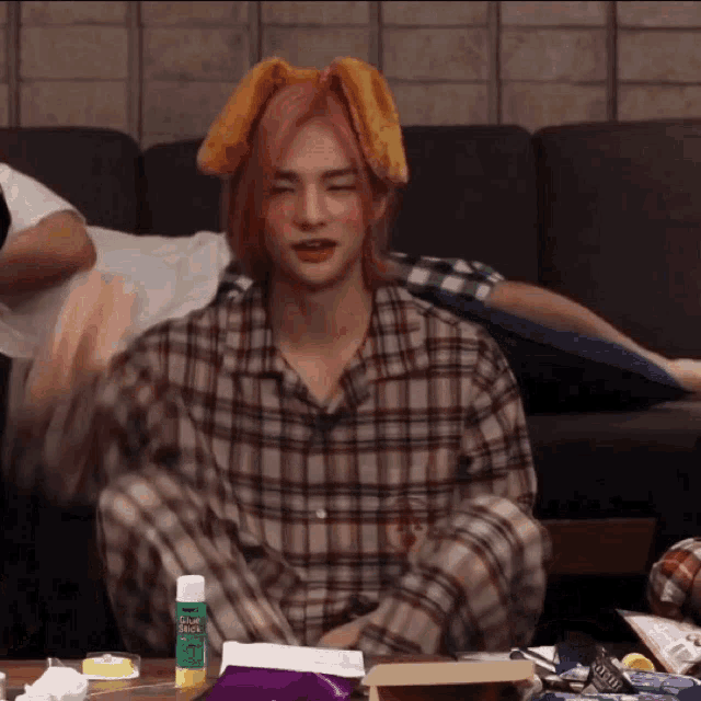 a man with pink hair is sitting on a couch wearing a plaid shirt and a towel on his head .