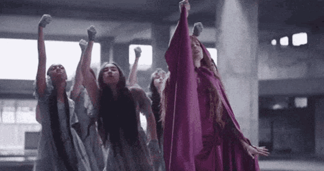 a group of women in purple dresses are standing in a room with their fists in the air .