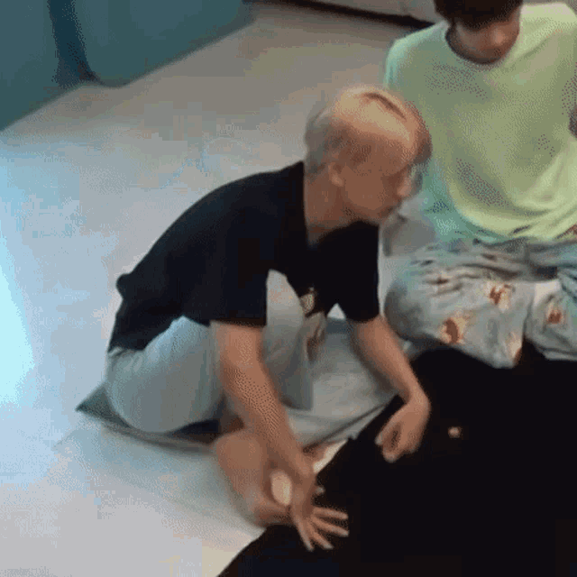 a man in a black shirt is kneeling on the floor while another man in a green shirt sits behind him .