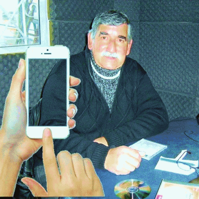 a man with a mustache is sitting at a table while a hand holds a cell phone in front of him