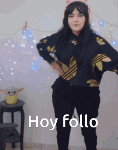 a woman wearing a black adidas sweater stands in front of a white wall with hoy follo written in white letters