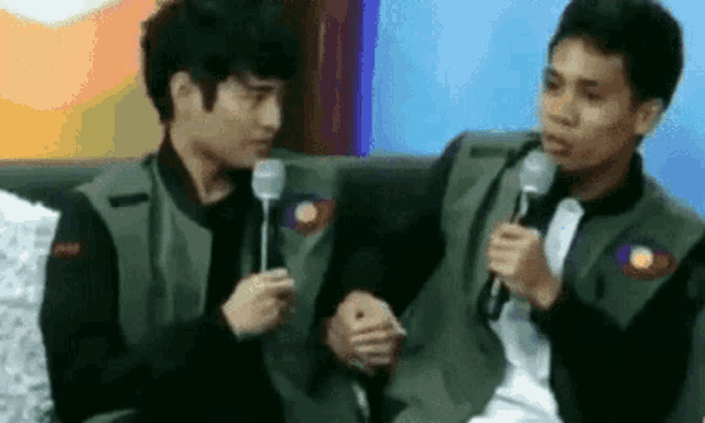two men are sitting next to each other on a couch holding hands and talking into microphones .