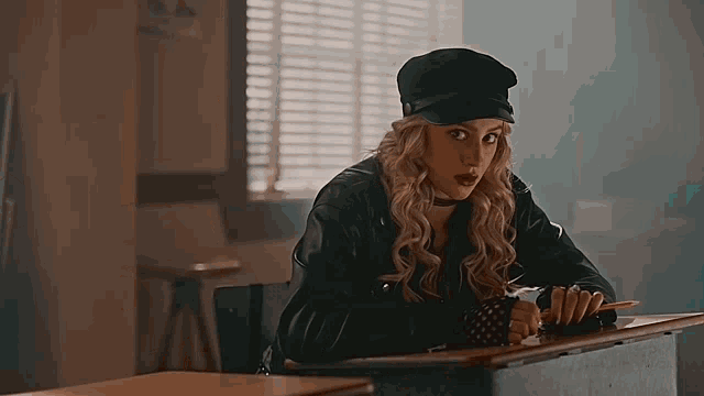 a woman wearing a hat and a leather jacket is sitting at a desk in a classroom .