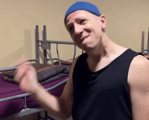 a man wearing a blue hat and a black tank top is standing in front of a bunk bed