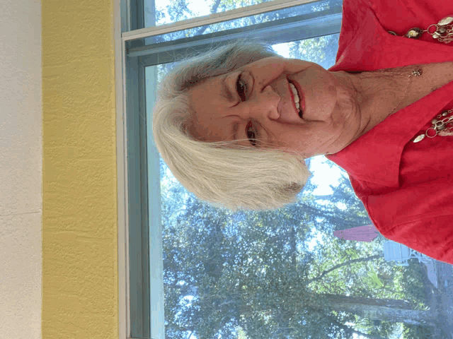 a woman wearing a red shirt and a necklace smiles in front of a window