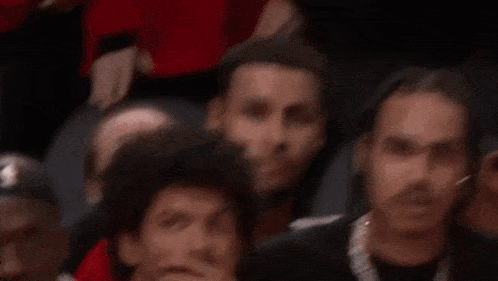 a group of men are sitting in a stadium watching a basketball game and one of them is wearing a red shirt .