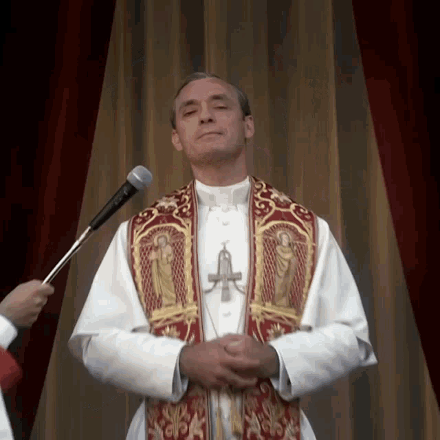 a priest stands in front of a microphone wearing a red and gold stole