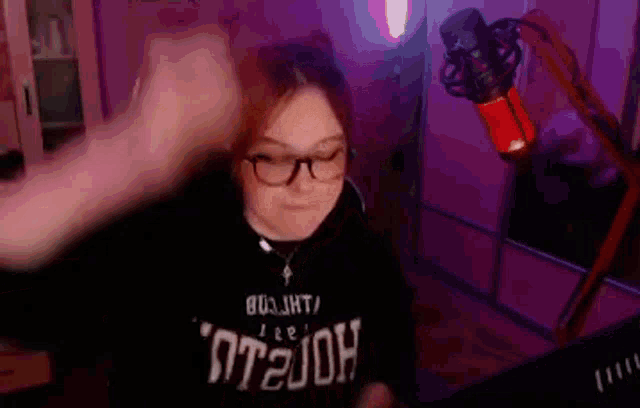 a woman wearing glasses and a black shirt is standing in front of a microphone in a room .