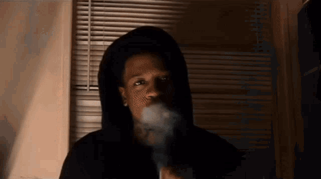 a man in a hooded sweatshirt is smoking a cigarette in front of a window .