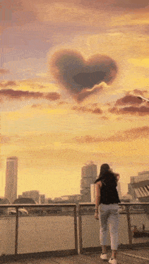 a woman stands on a bridge looking at a heart shaped cloud