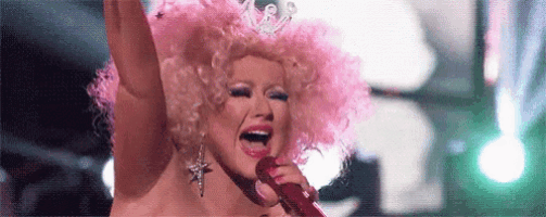 a woman in a pink wig is singing into a microphone with her arms in the air .