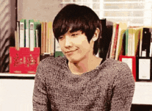 a young man is sitting in front of a bookshelf wearing a sweater .