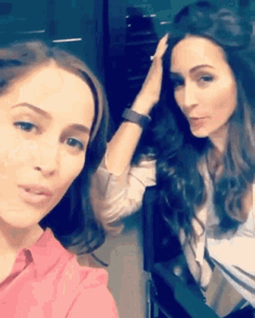 two women are standing next to each other in a room and one is holding her hair .