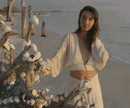 a woman in a white dress is standing next to a tree on the beach
